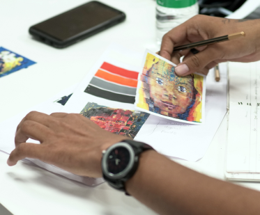 A person holds a pencil and pixelated face artwork over a notebook on a desk with a phone, water bottle, and printed images, suggesting creative work.