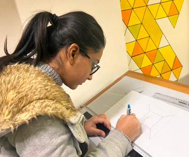 A person with glasses and a ponytail draws a geometric design with a blue pen and ruler at a desk, against a yellow and orange mural.