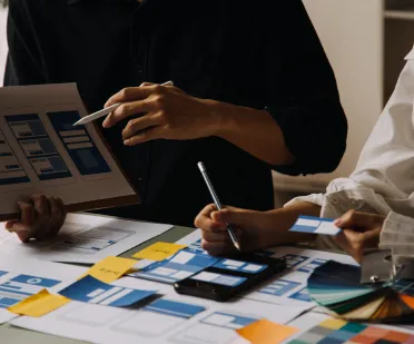 A group discusses design layouts on a tablet, with color swatches and papers scattered; one uses a stylus, another holds a pen.