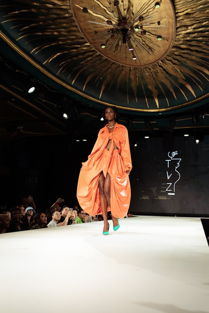 A model walks the runway in a bright orange outfit and green heels, under an ornate ceiling, with a "TVZ" logo on the black backdrop.