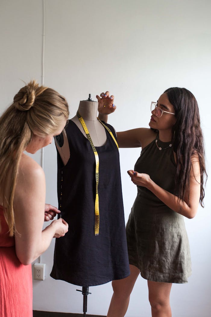 Two women adjust a black dress on a dress form. One in an orange dress, the other in green, with a measuring tape around its neck.