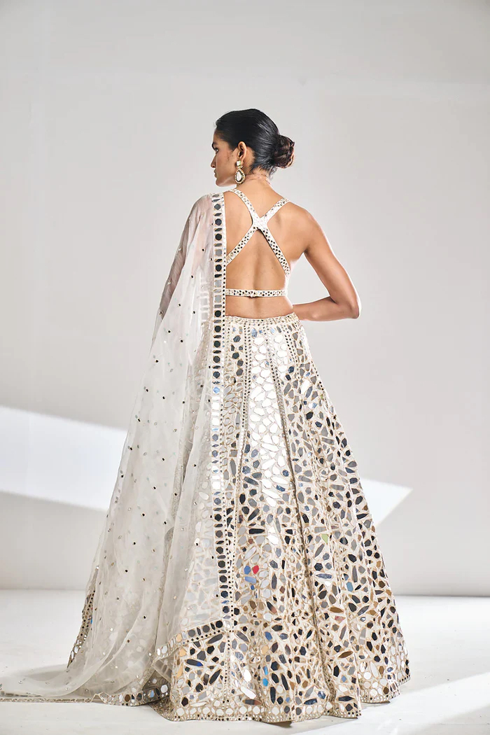 A woman in an ornate, backless gown with mirror work stands with her back to the camera, her hair in an elegant updo.