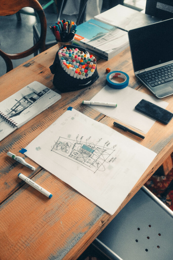 A cluttered desk holds architectural drawings, markers, a laptop, a smartphone, and blue tape, with sketches and a notebook on rustic wood.