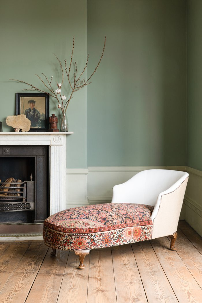 A serene room with green walls has a white marble fireplace with vases and a white chaise longue with a colorful cushion on a light wooden floor.