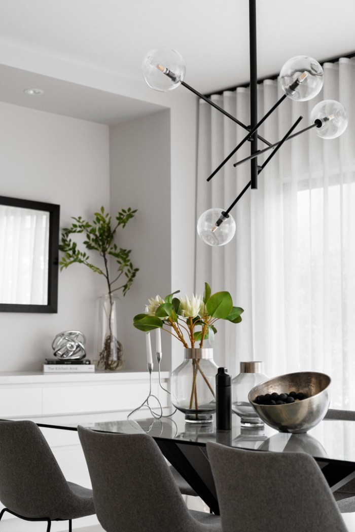 Modern dining room with a sleek glass table, gray chairs, globe chandelier, greenery vase, and stylish decor. White curtains add elegance.