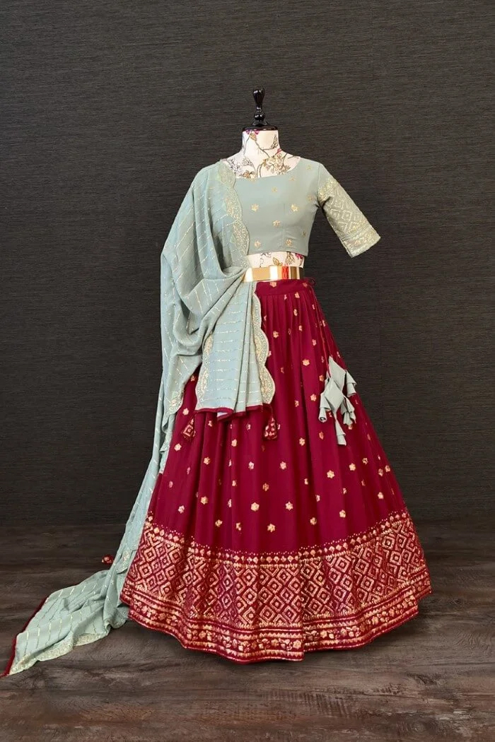 A mannequin showcases an elegant traditional Indian outfit: a light blue blouse, gold-accented dupatta, and maroon lehenga with gold embroidery.