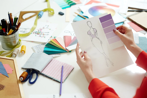 A person holds a fashion sketch and purple swatch at a cluttered table with colored pencils, fabric swatches, and scissors.