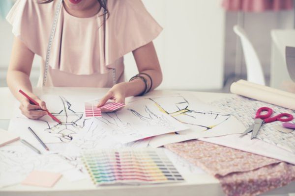 A person sketches fashion designs at a table with colored pencils, fabric swatches, scissors, and a measuring tape, wearing a light pink garment.