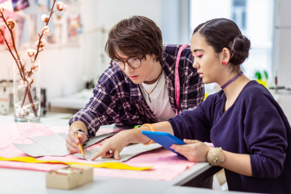 Two young people work at a table in a creative studio; one cuts fabric while the other points at a tablet, with tools nearby.