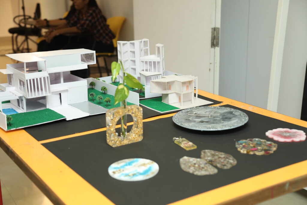 A table displays architectural models, a small plant, resin art, and a plate, with a person seated at another table in the background.