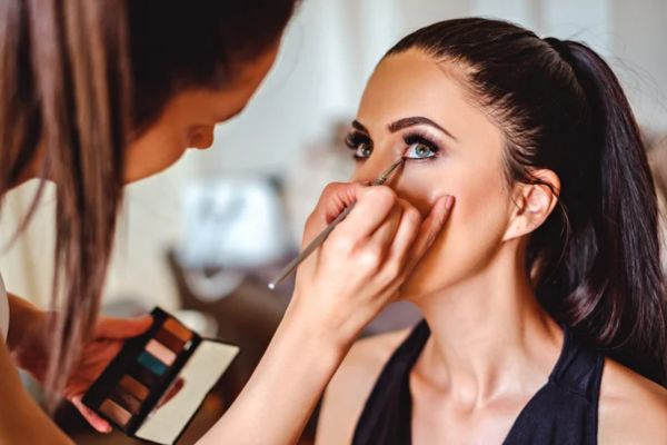 A makeup artist applies eyeshadow to a woman with straight dark hair tied back, who gazes upward while the artist holds a color palette.