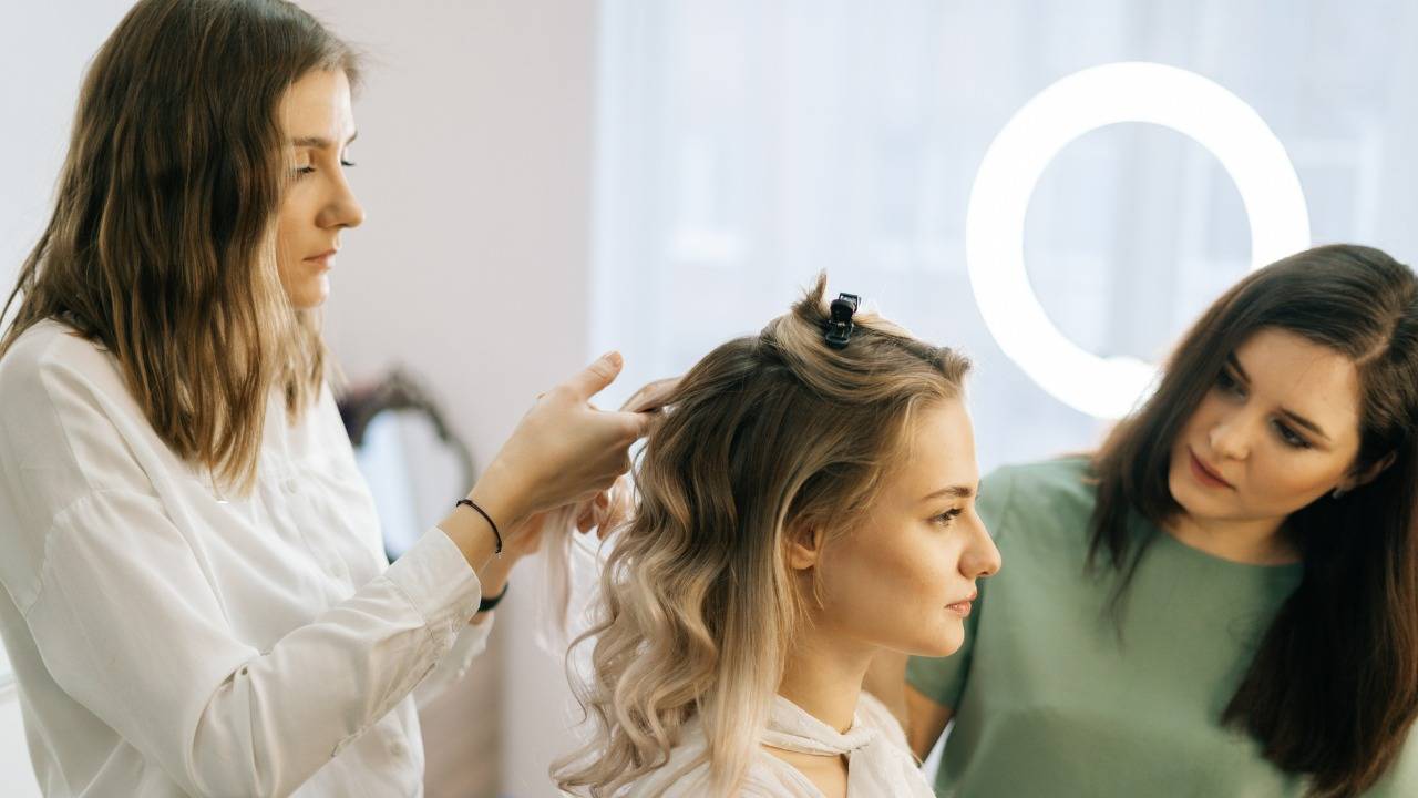 A blonde woman gets her hair styled with a curling iron by a brunette, while another brunette watches. A circular light is in the background.