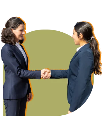 Two women in dark suits shake hands and smile in a professional setting, indicating a formal greeting or agreement against a solid background.