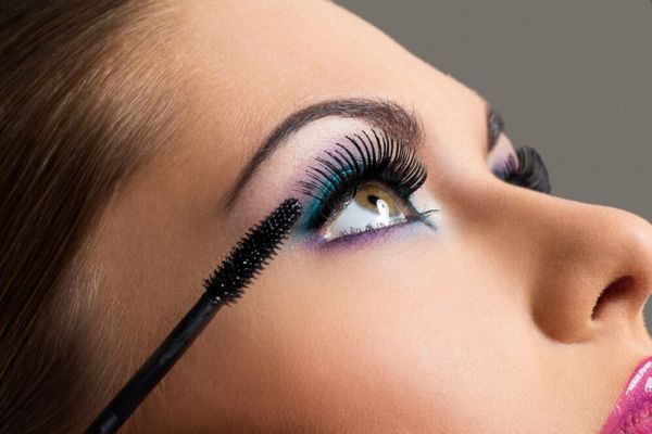 Close-up of a woman applying black mascara, with bold teal and purple eye makeup against a soft gray background.