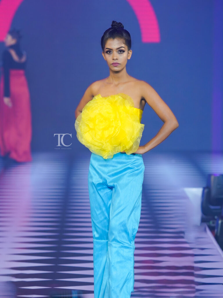 A model walks the runway in blue pants and a strapless top with a large yellow flower, alongside another model in red and black.