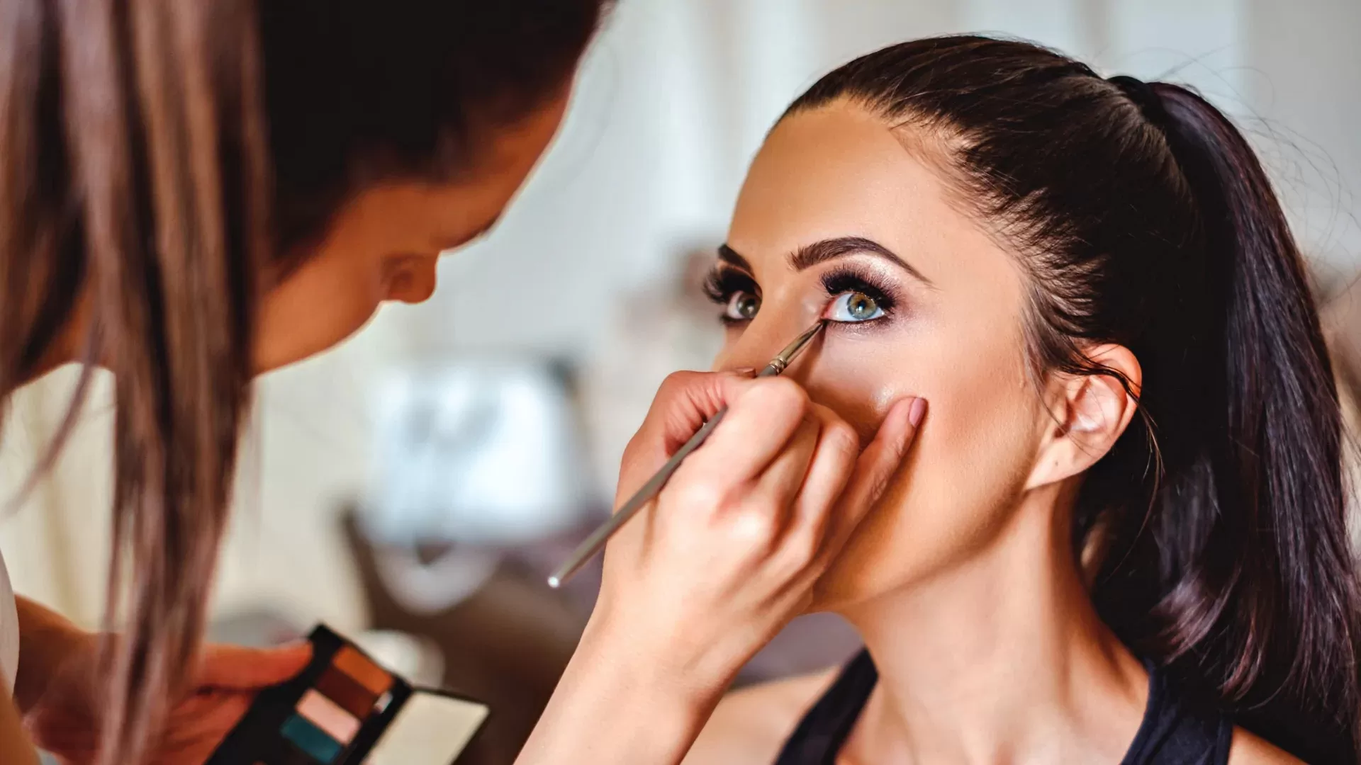 A person with long dark hair tied back has their eye makeup applied indoors by a makeup artist holding a brush and palette.