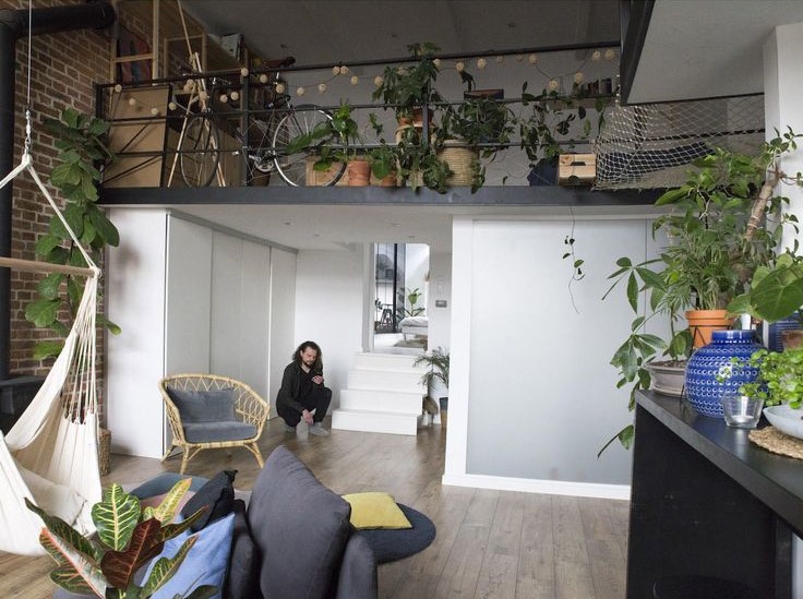 A person sits on a gray chair in a modern, plant-filled living space with wooden floors and a hammock. Stairs lead to a loft.