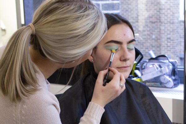 A blonde applies vibrant makeup to a seated person with closed eyes and teal eyeshadow in a room with a large window and ledge items.