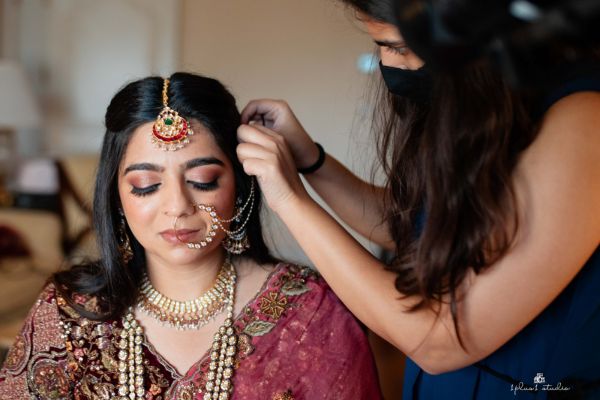 A bride in maroon attire and heavy jewelry closes her eyes as a woman adjusts her headpiece, showing intricate henna and makeup.