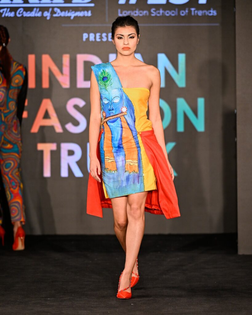 A model walks the runway in a vibrant off-shoulder dress with an abstract design and red draping, against a backdrop for "Indian Fashion Trunk."