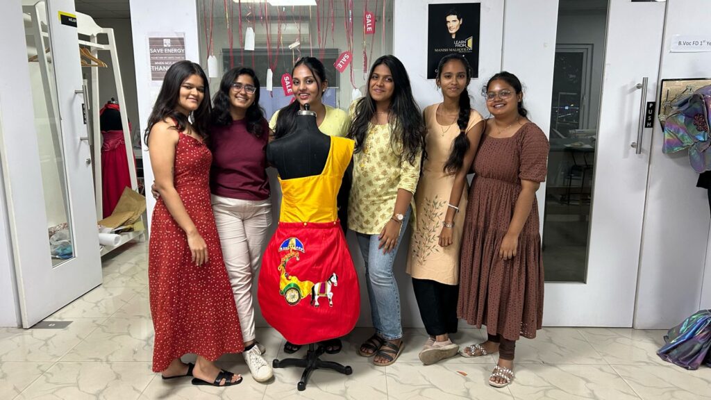 Six women smile beside a dress form in a bright yellow and red garment with colorful embroidery, in a well-lit room filled with clothing and decor.