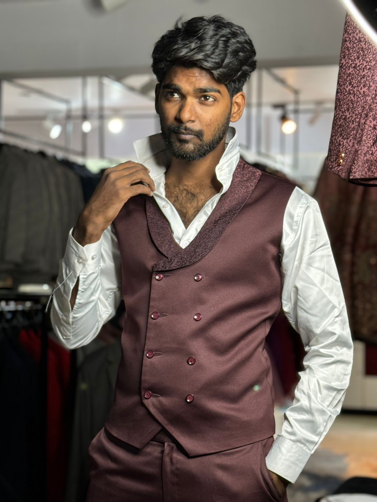 A man in a maroon tuxedo and white shirt stands confidently in a well-lit clothing store, holding his collar with one hand.