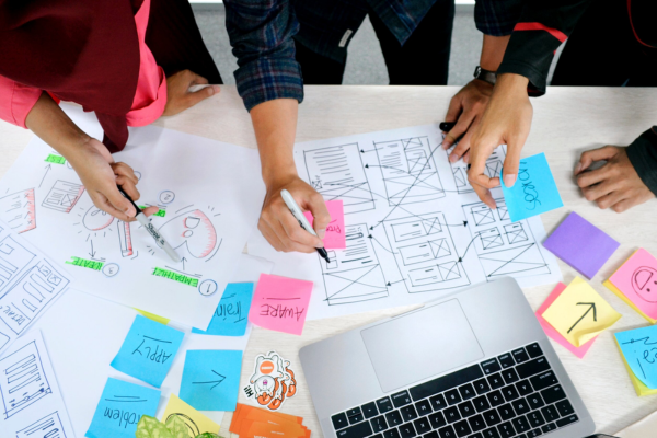 A group collaborates around a table with design sketches, wireframes, and sticky notes, actively brainstorming with a laptop nearby.