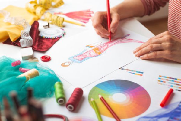 A person sketches a fashion design with colored pencils, surrounded by spools of thread, fabric swatches, and other design tools.