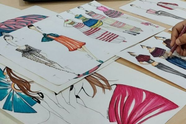 A person sketches fashion designs with colored markers, surrounded by vibrant illustrations of dresses and outfits on the table.