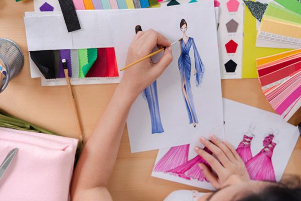 An artist's desk shows hands sketching a model in a blue dress, surrounded by colored fabrics, paint swatches, and design tools.