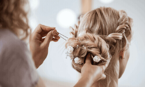 A person styles an intricate updo, securing it with a bobby pin. The hairstyle features twists and small white flowers for elegance.