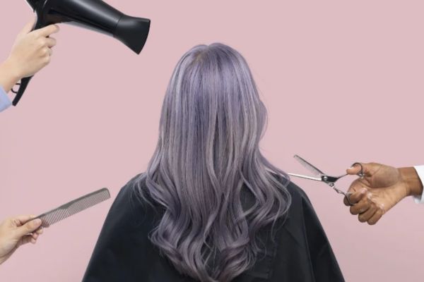 A person with long lavender hair sits draped in black, as hands hold a hairdryer, comb, and scissors around them in a salon setting.