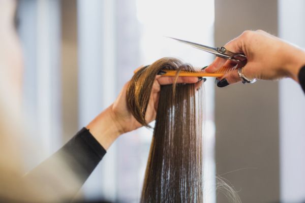 A hairstylist trims the ends of a client's straight brown hair, sectioned with a comb, against a softly blurred background.