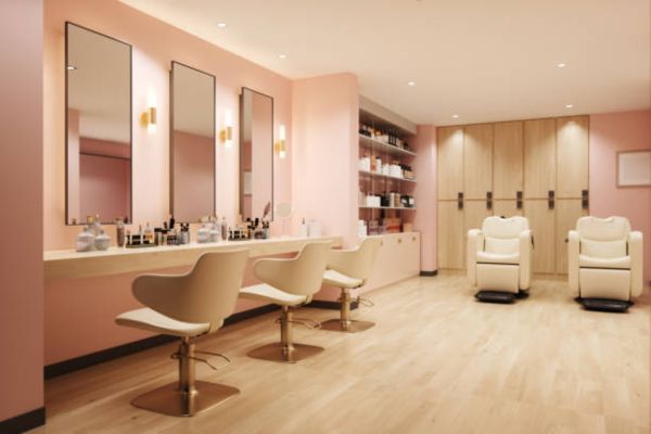 A brightly lit salon with pink walls has three beige chairs with gold bases in front of mirrors and shelves of products behind.