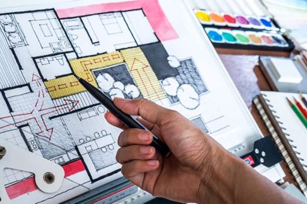 A hand holds a smartphone above a detailed architectural floor plan sketch, with drawing tools, colored pencils, and a notebook nearby.