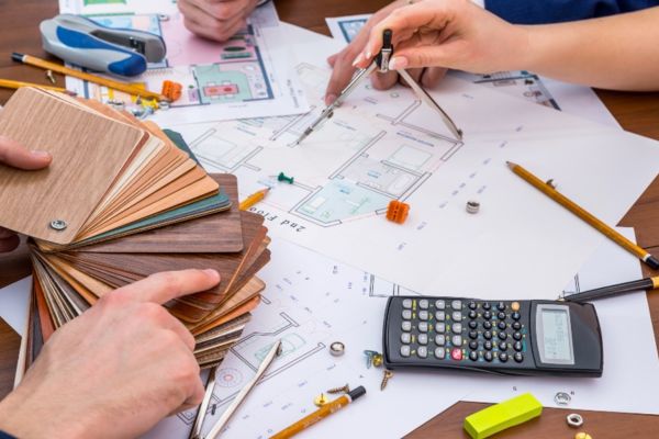 A cluttered desk has architectural blueprints, a calculator, compass, color swatches, and pencils, with hands pointing at them.