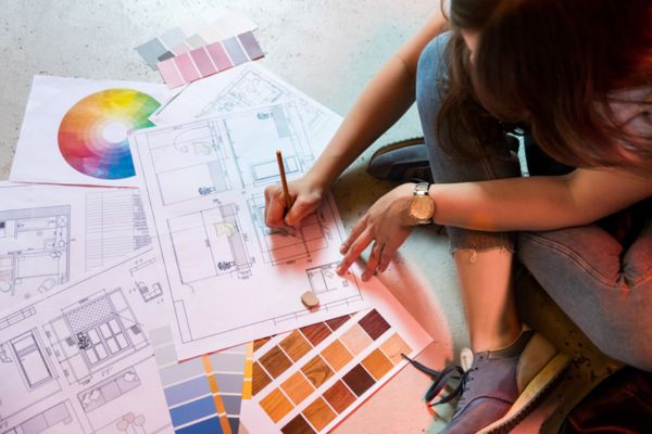 A person sits on the floor, sketching with a pencil amid architectural blueprints, color swatches, and design plans.