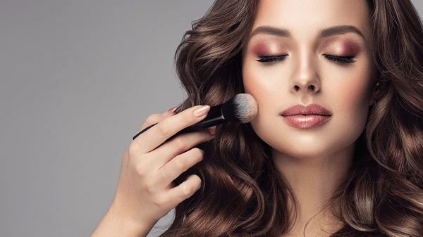 A woman with wavy brown hair applies cheek makeup with a brush, eyes closed and serene, in warm smoky tones against a gradient background.