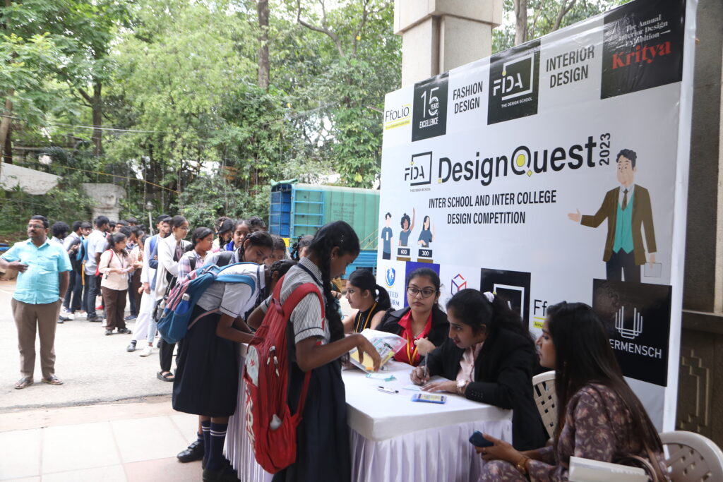 Students line up at a registration table for "DesignQuest 2023," staffed by individuals under a banner with logos.