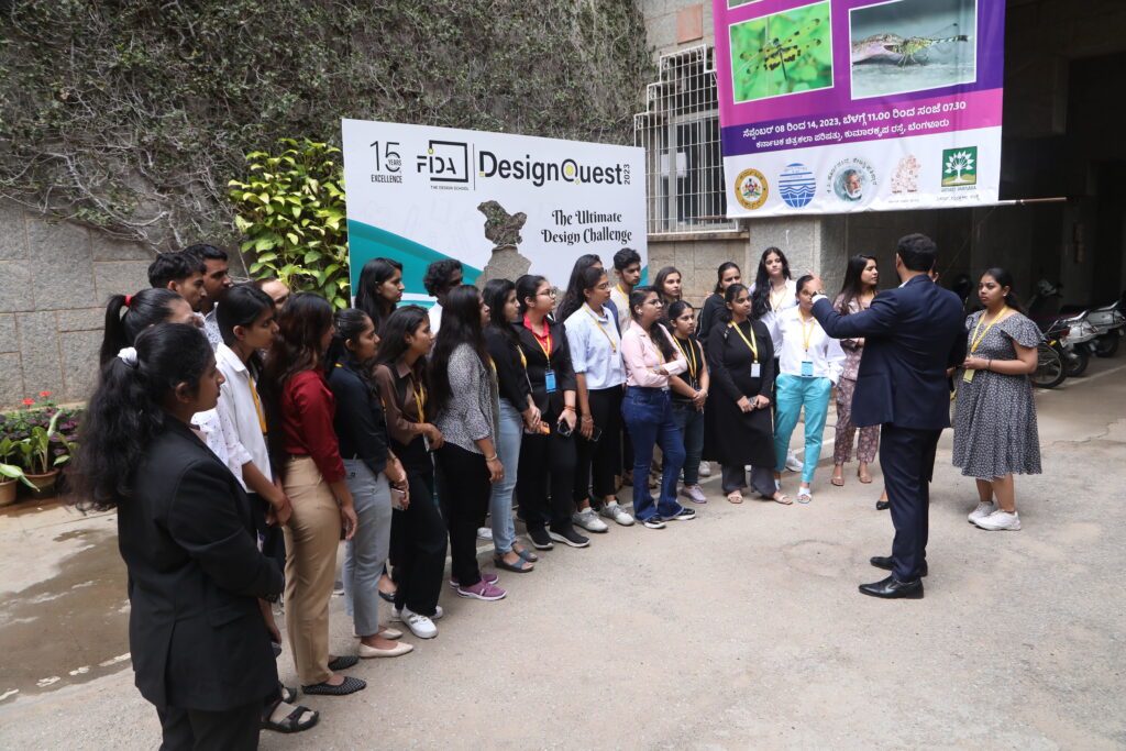 A group listens to a speaker outside, with "DesignQuest" banners and signage behind them, set against greenery and a building.