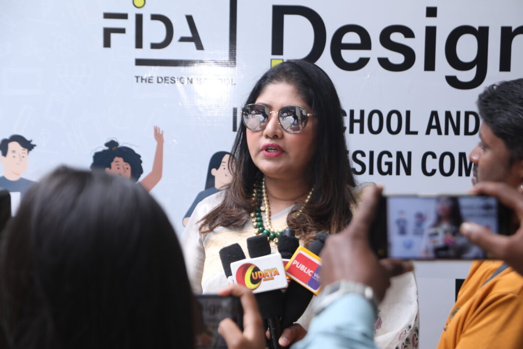 A woman in sunglasses speaks into multiple microphones against a "FIDA" and "Design" backdrop while reporters record her.