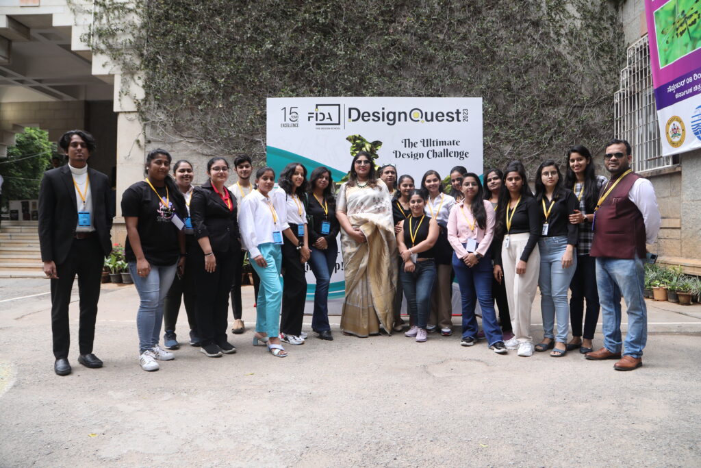 A group stands in front of a "DesignQuest" banner for "The Ultimate Design Challenge." Most wear casual attire; one woman is in a sari.