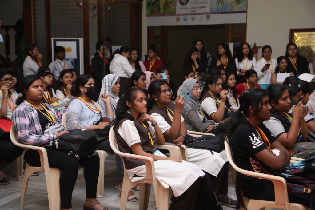 A group of students, mainly young women, listens attentively to a presentation, some taking notes or photos, in an engaging atmosphere.