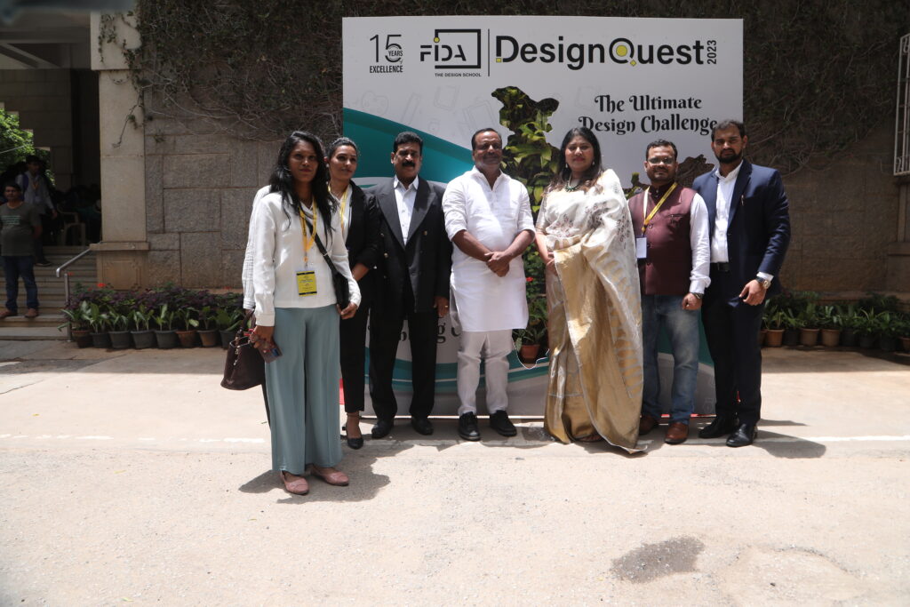 A group of three women and four men in formal attire stands by a "DesignQuest 2023" sign, with plants and a vine-covered building behind.