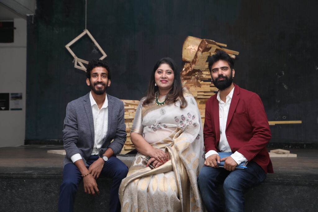 Three people smile against a dark geometric backdrop: a man in a gray blazer, a woman in a saree, and a man in a red blazer.