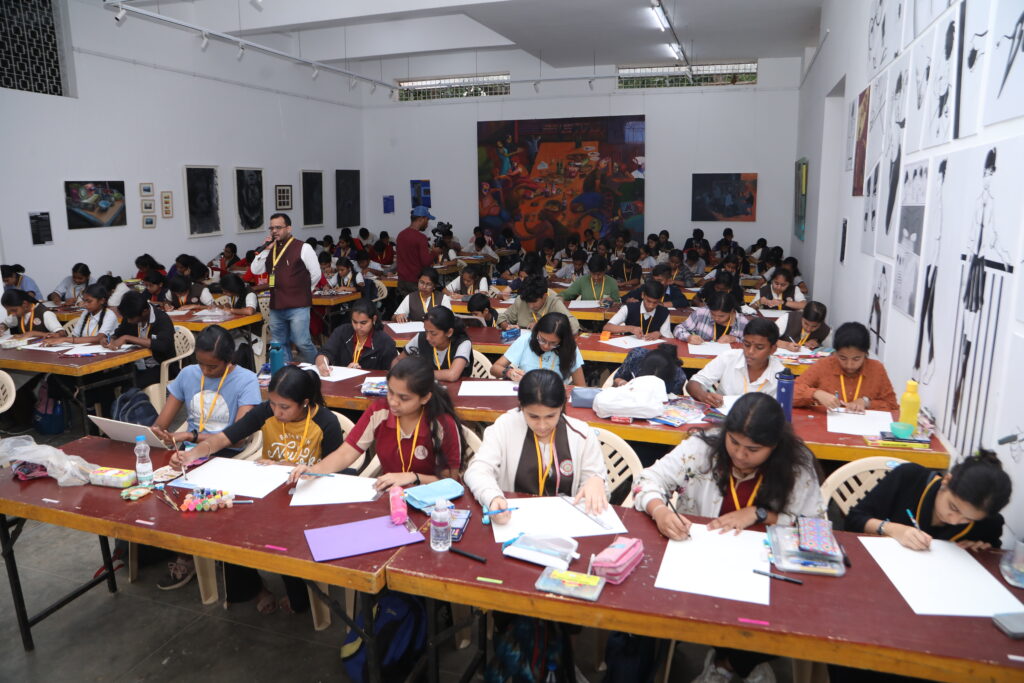 A large classroom has students at long tables drawing, with artworks on the walls and an instructor in a maroon sweater observing.