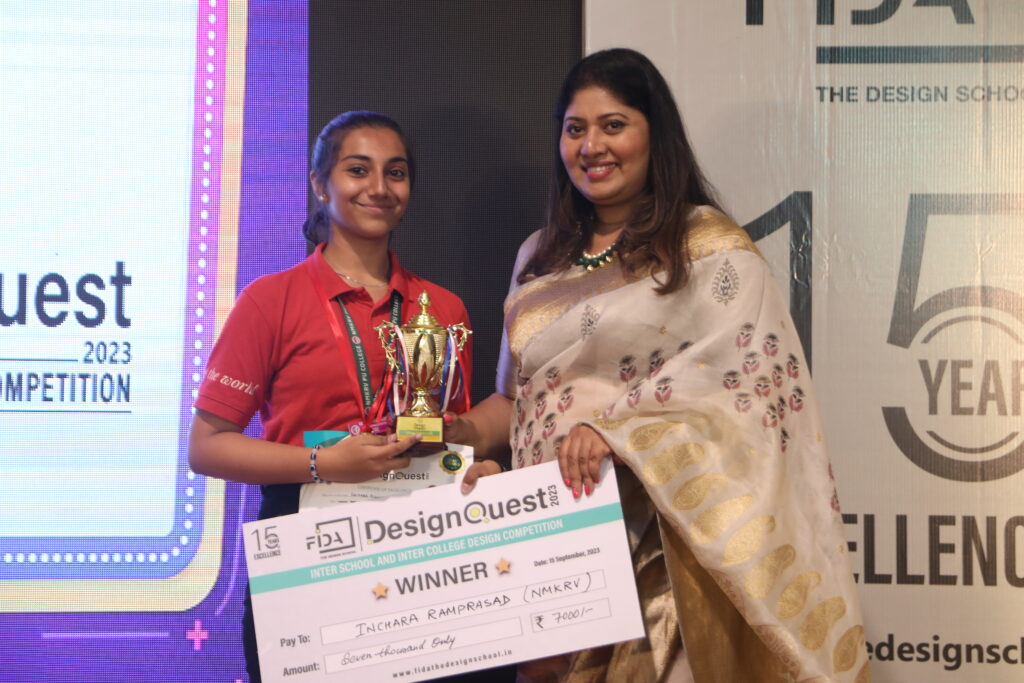 Two smiling women stand on stage; one in a red shirt holds a trophy and check, while the other wears a traditional outfit.