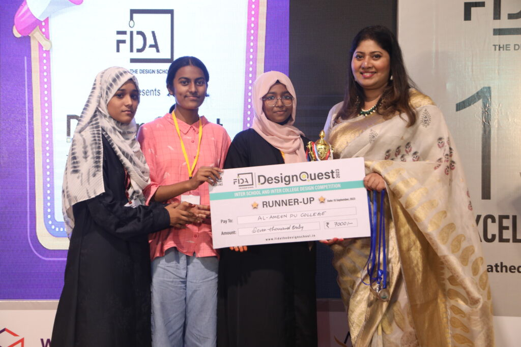 Four women hold a runner-up cheque from FIDA Design Quest for Al-Ameen PU College. The sari-clad woman smiles with a microphone.