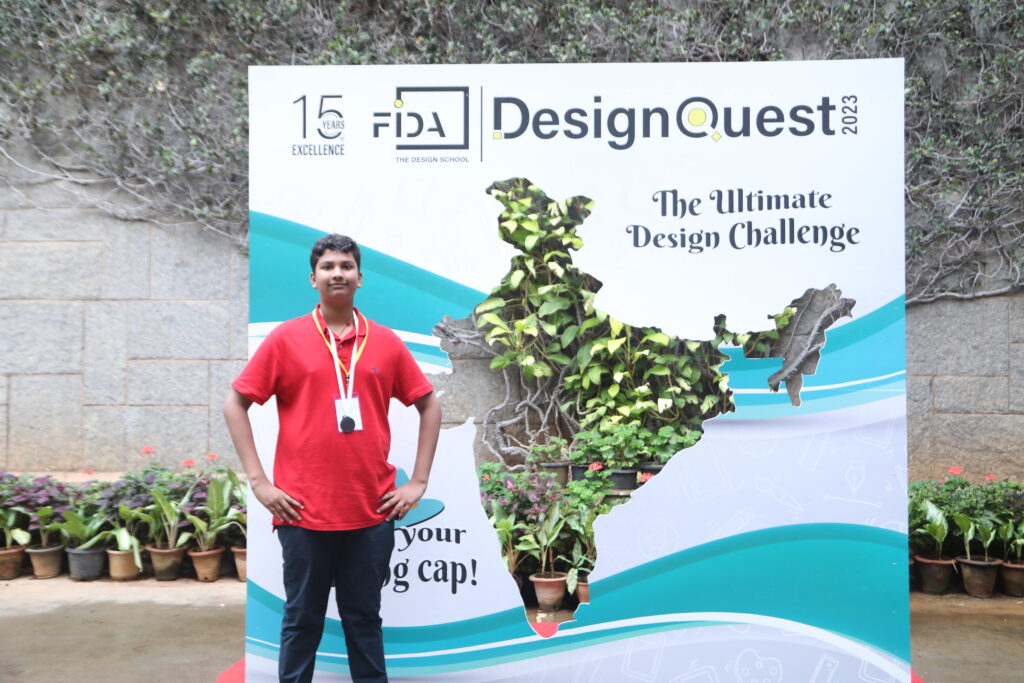 A young person in a red shirt stands before a green plant map of India backdrop for "DesignQuest 2023," with potted plants behind.