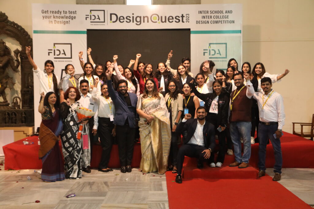 A group on stage in front of a "DesignQuest" backdrop smiles and raises their hands, holding certificates and trophies from a school design competition.